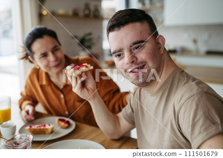 Young man with Down syndrome having breakfast with his mother at home. Morning routine for man with Down syndrome. Young man with Down syndrome having breakfast with his mother at home. Morning routine for man with Down syndrome. 111501679