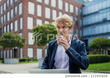 Businessman, freelancer or manager working outdoors in city park. Man with earbuds drinking coffee and listening music. Concept of working remotely. Businessman, freelancer or manager working outdoors in city park. Man with earbuds drinking coffee and listening music. Concept of working remotely. 111501828