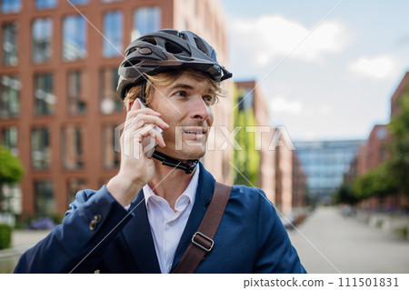Handsome man commuting through the city by bike, making call with smartphone. Male city commuter traveling from work by bike after a long workday. Handsome man commuting through the city by bike, making call with smartphone. Male city commuter traveling from work by bike after a long workday. 111501831