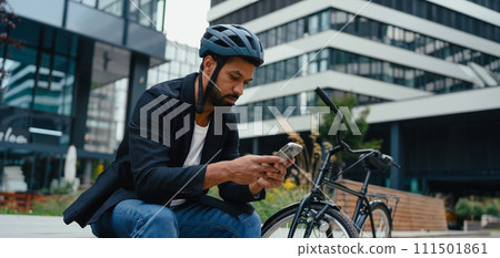 City commuter, sitting on bench, scrolling on smartphone. Businessman traveling from work to home on bike. City lifestyle. 111501861