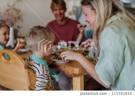 Family eating breakfast together in home kitchen. Healthy breakfast or snack before school and work. Mother helping youngest son. Family eating breakfast together in home kitchen. Healthy breakfast or snack before school and work. Mother helping youngest son. 111501916
