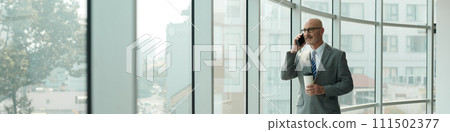 Header of confident mature male solopreneur in grey suit speaking on smartphone while looking through large window in office center Header of confident mature male solopreneur in grey suit speaking on smartphone while looking through large window in office center 111502377