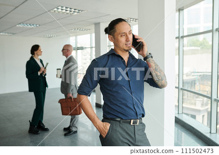 Young Asian male solopreneur in shirt and pants speaking on mobile phone while standing in front of camera in office center Young Asian male solopreneur in shirt and pants speaking on mobile phone while standing in front of camera in office center 111502473