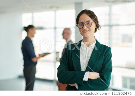 Young successful female chief executive officer in eyeglasses and formalwear looking at camera with smile against two colleagues Young successful female chief executive officer in eyeglasses and formalwear looking at camera with smile against two colleagues 111502475