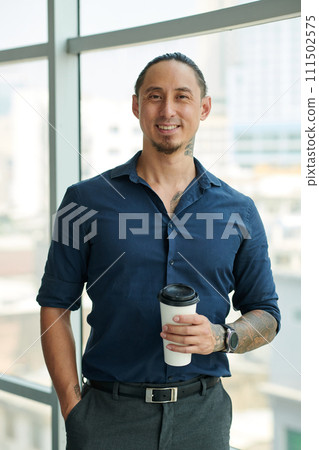 Young male employee or solopreneur in dark blue shirt looking at camera with smile at coffee break while standing against large window Young male employee or solopreneur in dark blue shirt looking at camera with smile at coffee break while standing against large window 111502575