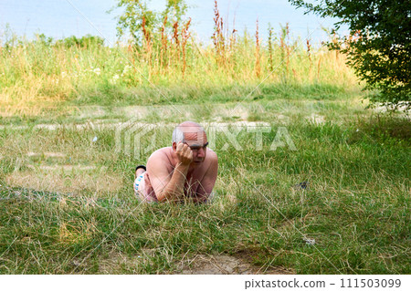 An elderly man is resting on a grass in a lawn near a summer river 111503099