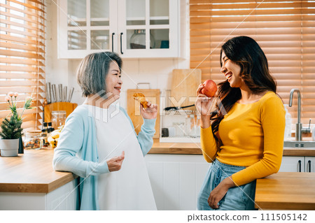 An elderly mother and her adult daughter, a young woman in an apron, bond over food in the kitchen. Their cheerful smiles highlight the happiness and fun of family togetherness at home. An elderly mother and her adult daughter, a young woman in an apron, bond over food in the kitchen. Their cheerful smiles highlight the happiness and fun of family togetherness at home. 111505142