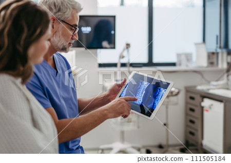 Doctor showing female patient MRI scan on tablet, discussing test result in emergency room. 111505184