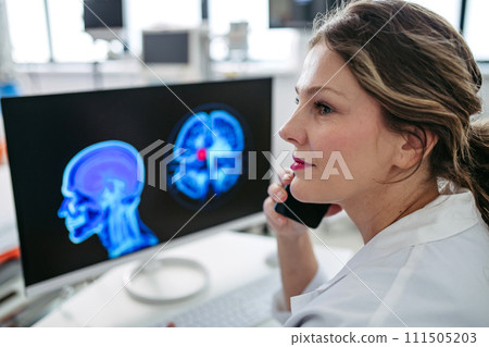 Female doctor working on computer in doctor's office, looking at MRI scan, phone calling test results to patient. Doctor consulting scan with other doctors. 111505203