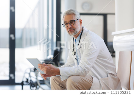 Portrait of smiling doctor sitting in hospital corridor. Handsome doctor with gray hair wearing white coat, stethoscope around neck standing in modern private clinic, looking at camera. Portrait of smiling doctor sitting in hospital corridor. Handsome doctor with gray hair wearing white coat, stethoscope around neck standing in modern private clinic, looking at camera. 111505221