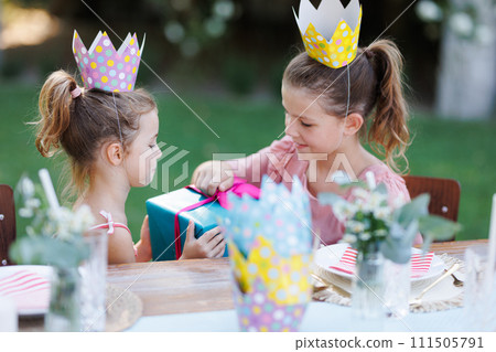 Little friends at birthday party outdoors at the garden. Cute girls with paper crowns opening birthday gift. Birthday garden party for children. Little friends at birthday party outdoors at the garden. Cute girls with paper crowns opening birthday gift. Birthday garden party for children. 111505791