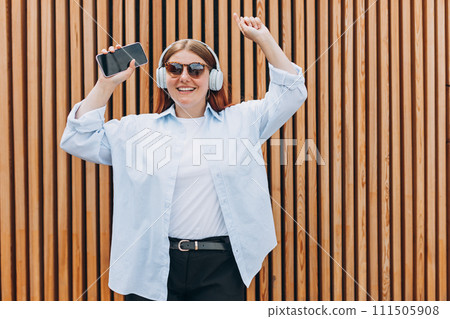 Closeup portrait of a dancing young woman listening to music through wireless earphones on wooden rustic background. Music lover enjoying music. Closeup portrait of a dancing young woman listening to music through wireless earphones on wooden rustic background. Music lover enjoying music. 111505908