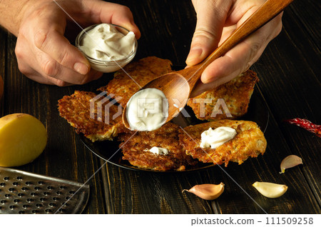 Close-up of a spoon with sour cream in the hand of a chef. Preparing a breakfast dish with potato pancakes and sour cream. Restaurant food serving concept Close-up of a spoon with sour cream in the hand of a chef. Preparing a breakfast dish with potato pancakes and sour cream. Restaurant food serving concept 111509258