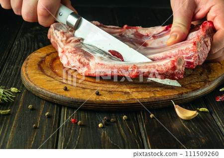 The cook cuts raw lamb ribs with a knife on a wooden board. The concept of the process of preparing a meat dish or kebab. Asian cuisine The cook cuts raw lamb ribs with a knife on a wooden board. The concept of the process of preparing a meat dish or kebab. Asian cuisine 111509260