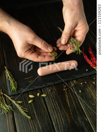 The chef is preparing milk sausage with rosemary on the kitchen table. A cook adds rosemary to a raw sausage by hand before baking. Free space for advertising The chef is preparing milk sausage with rosemary on the kitchen table. A cook adds rosemary to a raw sausage by hand before baking. Free space for advertising 111509263