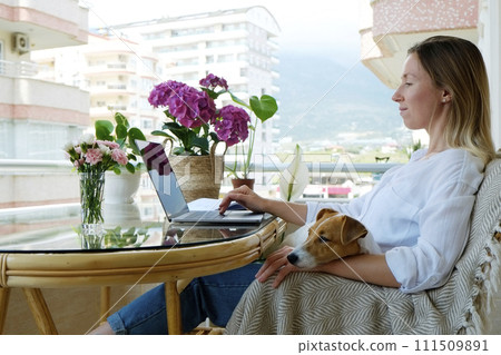 Young beautiful woman sitting on the armchair freelancing on a balcony. Female working from comfort of her home with jack russell terrier puppy. Interior, background, close up, copy space. Young beautiful woman sitting on the armchair freelancing on a balcony. Female working from comfort of her home with jack russell terrier puppy. Interior, background, close up, copy space. 111509891