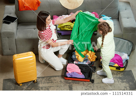 Mother with little daughter choosing and packaging clothes to big suit cases in living room at home. Joyful pre-travel mood. Preparing for family trip. 111514331