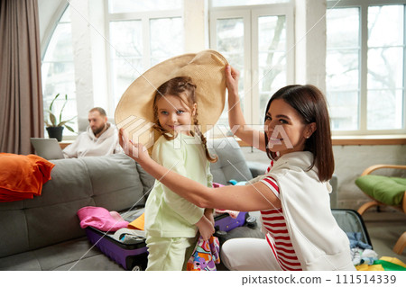 Little daughter helps her mother choosing clothes for travel and have fun with mother tries to girl to hat in living room at home. Little daughter helps her mother choosing clothes for travel and have fun with mother tries to girl to hat in living room at home. 111514339
