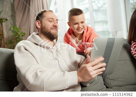 portrait of man, dad shows child, his son, something on tablet while sitting on couch in living room at home. portrait of man, dad shows child, his son, something on tablet while sitting on couch in living room at home. 111514362