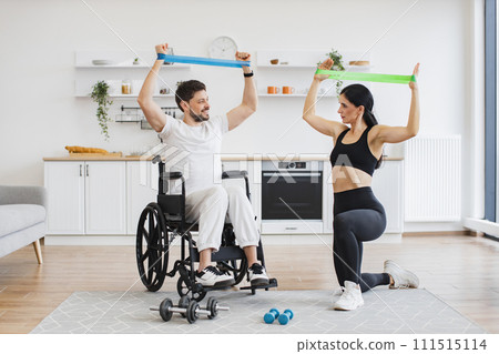 Wife helping mature man in wheelchair lifting arms with stretching bands. 111515114