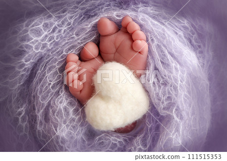 The tiny foot of a newborn baby. Soft feet of a new born in a lilac, purple wool blanket. Close up of toes, heels and feet of a newborn. Knitted white heart in the legs of a baby. Macro photography. 111515353