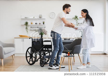 Caucasian woman helping patient with disability to stand from wheelchair. Caucasian woman helping patient with disability to stand from wheelchair. 111515938