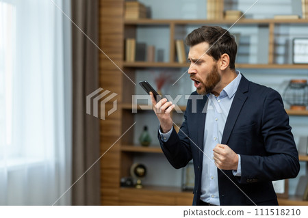 Angry young male businessman standing in the office in a suit and emotionally talking on the phone. Holds a mobile phone and shouts into the loudspeaker, solves questions and problems. 111518210