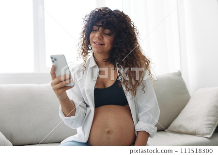 Pregnant woman blogger sits on the couch at home and takes pictures of herself on the phone, selfie and video call, online doctor consultation, pregnancy management, freelance work smiling 111518300