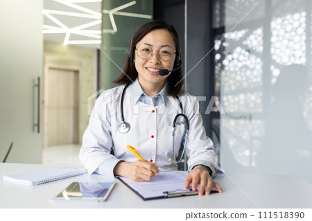 Portrait of a young Asian female doctor working in the office at a table with documents. Sits in a headset, consults and talks online. He looks at the camera with a smile. 111518390