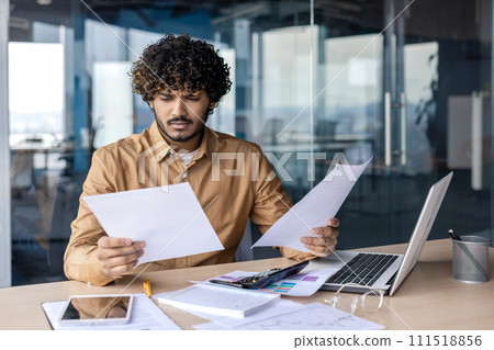 Worried young Indian male businessman working in office with laptop and documents, holding papers and looking disappointed. 111518856