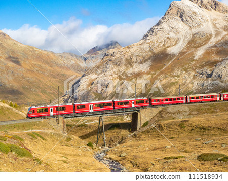 Bernina, Switzerland - October 15th 2023: Red train of Rhaetian railway passing a bridge in alpine terrain. Bernina, Switzerland - October 15th 2023: Red train of Rhaetian railway passing a bridge in alpine terrain. 111518934