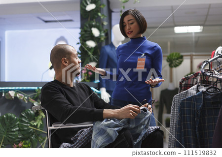 Arab man shopper with physical disability receiving assistance in clothing store from asian woman consultant. Shopping mall boutique client in wheelchair talking with seller 111519832