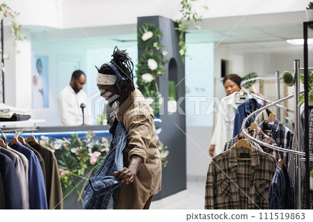 Cheerful african american man in headphones holding shirt on hanger while shopping in clothing store. Customer in good mood listening to music while choosing new outfit in fashion boutique Cheerful african american man in headphones holding shirt on hanger while shopping in clothing store. Customer in good mood listening to music while choosing new outfit in fashion boutique 111519863