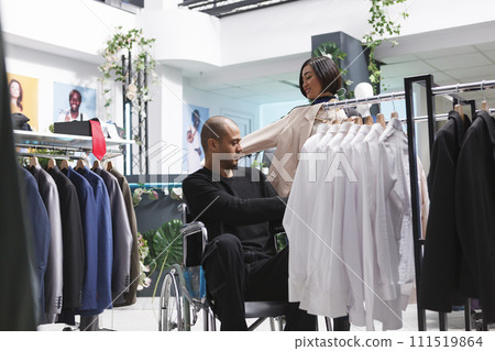 Mall asian woman seller helping arab man customer in wheelchair to choose female formal jacket. Clothing store consultant assisting client with disability in selecting stylish apparel Mall asian woman seller helping arab man customer in wheelchair to choose female formal jacket. Clothing store consultant assisting client with disability in selecting stylish apparel 111519864