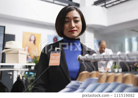 Asian woman seller working in clothing store, managing formal apparel inventory and checking shits on display rack. Shopping center employee organizing menswear garment in boutique Asian woman seller working in clothing store, managing formal apparel inventory and checking shits on display rack. Shopping center employee organizing menswear garment in boutique 111519882