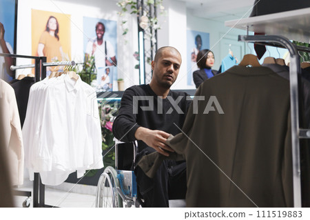 Arab buyer in wheelchair exploring formal menswear rack, seeking outfit in fashion boutique. Shopping mall clothing store client with physical disability selecting stylish garment Arab buyer in wheelchair exploring formal menswear rack, seeking outfit in fashion boutique. Shopping mall clothing store client with physical disability selecting stylish garment 111519883