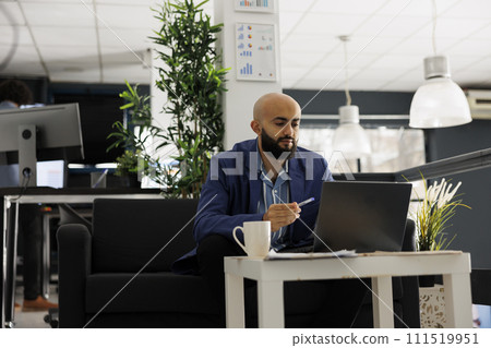 Start up business employee analyzing data on laptop in open space modern office. Arab young man entrepreneur doing marketing research while sitting on couch in coworking space 111519951