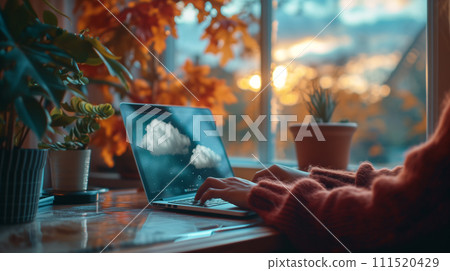A person works on a laptop with a cloud-themed screen, sitting comfortably at a home workstation by a window with autumn leaves outside. A person works on a laptop with a cloud-themed screen, sitting comfortably at a home workstation by a window with autumn leaves outside. 111520429