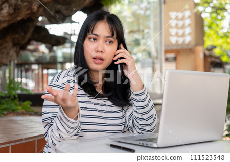 A serious Asian woman is talking on the phone with someone while working remotely at a cafe. A serious Asian woman is talking on the phone with someone while working remotely at a cafe. 111523548