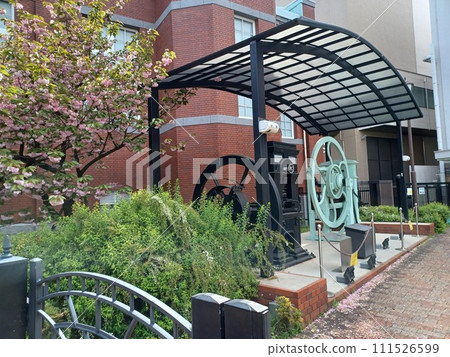In front of a brown building, cherry blossom petals gather at the feet of a machine displayed outdoors. Double cherry blossoms on the left 111526599