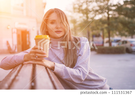 Woman drinks from cup on wooden bench. She is wearing a white shirt enjoying her beverage. The bench is located in a park setting, with trees in the background. Woman drinks from cup on wooden bench. She is wearing a white shirt enjoying her beverage. The bench is located in a park setting, with trees in the background. 111531367