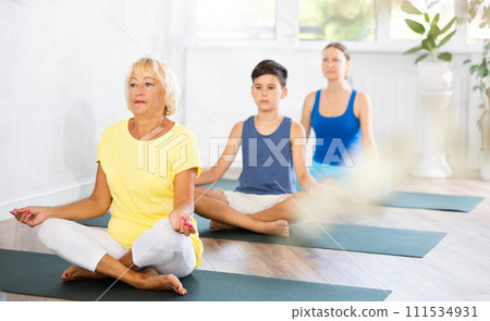 Old woman practicing half lotus pose of yoga in light fitness room Old woman practicing half lotus pose of yoga in light fitness room 111534931