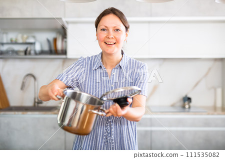 Near dining table in kitchen young woman wife standing with stainless saucepan Near dining table in kitchen young woman wife standing with stainless saucepan 111535082