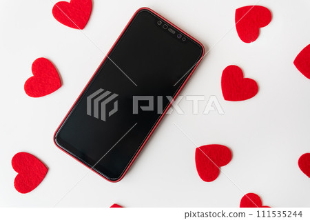 Smartphone surrounded by red felt hearts on a white background. Place for an inscription. Smartphone surrounded by red felt hearts on a white background. Place for an inscription. 111535244