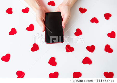 Hands holding a smartphone with scattered red hearts on a white background. Mockup, place for inscription. Hands holding a smartphone with scattered red hearts on a white background. Mockup, place for inscription. 111535245