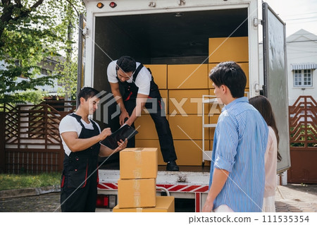 Professional movers assist a satisfied newlywed couple who sign the delivery checklist after furniture upload. Teamwork ensures a happy customer relocation. Moving Day Concept 111535354