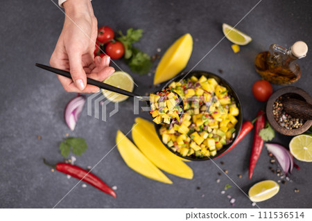 Freshly made mango salsa in a black ceramic bowl on a table with ingredients 111536514
