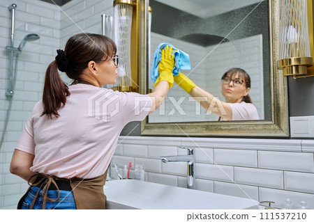 Middle-aged woman in an apron cleaning mirror in bathroom Middle-aged woman in an apron cleaning mirror in bathroom 111537210