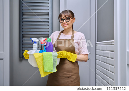 Middle-aged woman in gloves apron with basin of detergents in bathroom Middle-aged woman in gloves apron with basin of detergents in bathroom 111537212