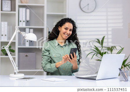 Professional hispanic woman working in a bright office space with a laptop and mobile phone, displaying efficiency and multitasking skills. Professional hispanic woman working in a bright office space with a laptop and mobile phone, displaying efficiency and multitasking skills. 111537451